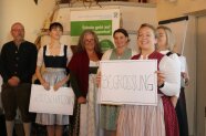 Frau hält Schild mit "Begrüßung" vor sich, Gruppe an Personen steht im Hintergrund